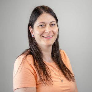 A woman with long brown hair wearing an orange shirt smiles brightly, radiating joy and positivity in her expression.
