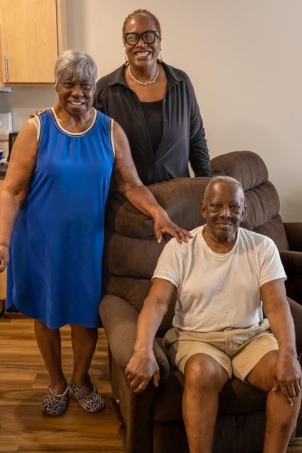 Two woman stand next to a man sitting in a brown arm chair