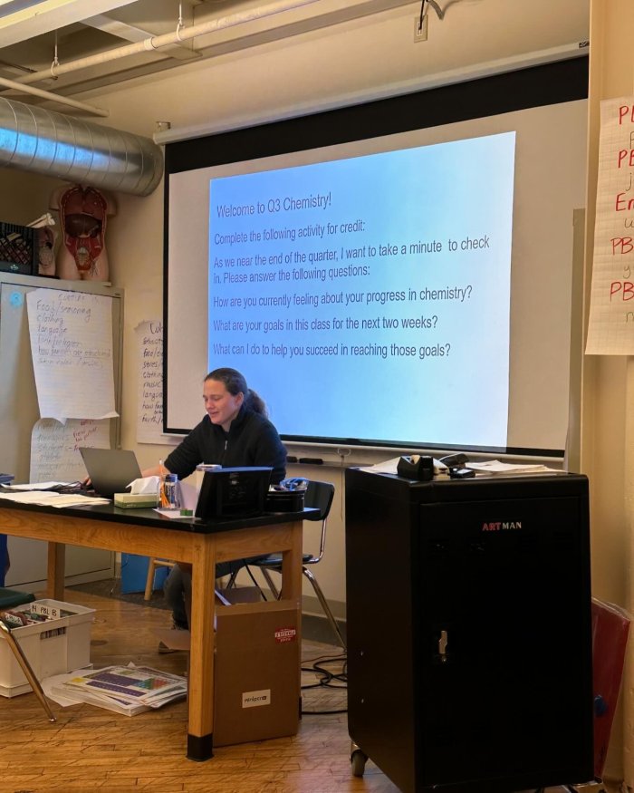 a woman sitting at a desk in front of a projector teaching