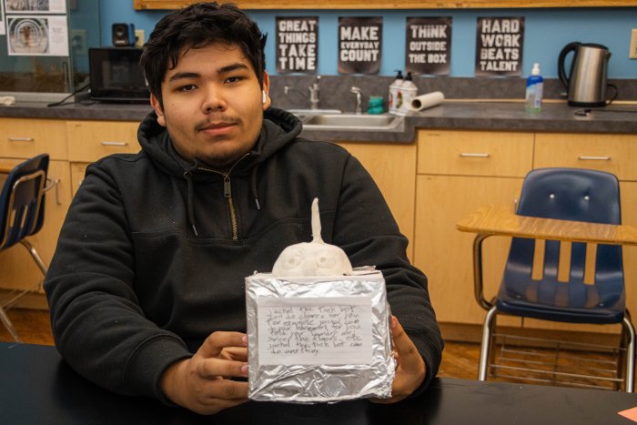 A student sitting at a desk holding a model of his invention 