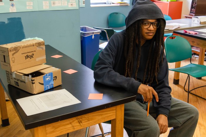 A student sitting at a desk next to a model of his invention made from cardboard boxes