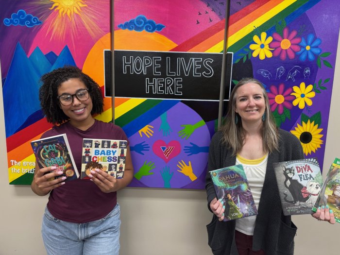 Two woman stand in front of a colorful mural holding up books smiling