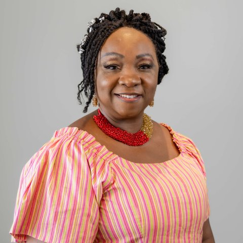 A woman with braided hair adorned with beads is wearing a pink and yellow striped dress, a red beaded necklace, and gold earrings, smiling against a plain background.