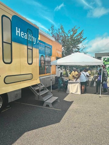 People gathered at a table under a tent next to a mobile clinic parked outside on a sunny day