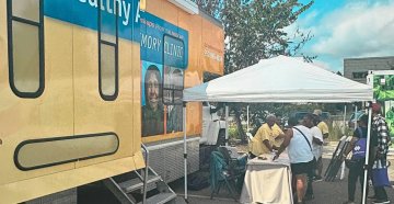 People gathered at a table under a tent next to a mobile clinic parked outside on a sunny day