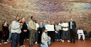 A group of diverse individuals standing together in front of a brick wall posing for a photo with two women in the center holding a giant check