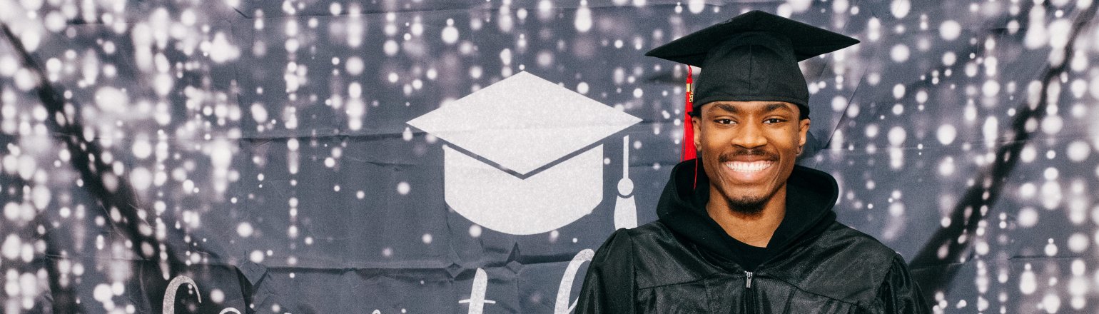 A young man smiling in a cap and gown in front of a Congratulations Class of 2025 banner