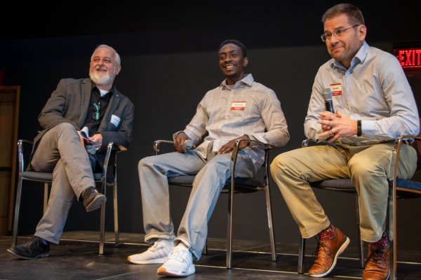 three men sitting in chairs on a stage holding microphones laughing