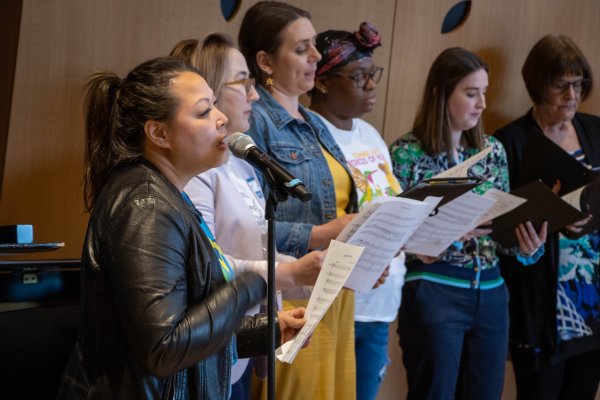 A women standing at a microphone singing a solo next to a choir of woman