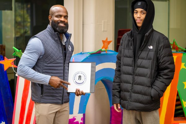 Two men stand together holding a Warrior Academy Binder, with bright, colorful letters in the background.