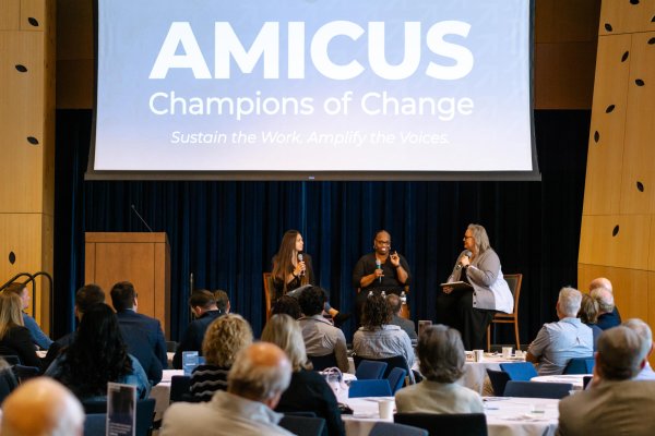 three women sitting in chairs on a stage at the front center of a room of people seated at tables