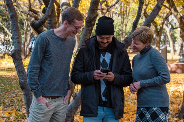 Fong showing Hans and Laura a photo on his phone while outside standing in front of a tree